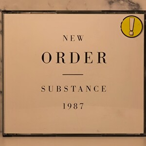 Peut inclure: Un boîtier de CD avec une couverture crème portant le nom du groupe "NEW ORDER" au-dessus du mot "SUBSTANCE" et l'année "1987". Un autocollant jaune avec un point d'exclamation est dans le coin supérieur droit. Le boîtier est dans un étui en plastique transparent.