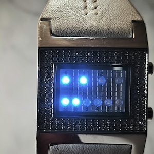 May include: A digital wristwatch with a rectangular face and a light grey leather strap. The watch face is black, with a grid of blue LED lights displaying the time. The bezel is black and encrusted with small, dark stones.