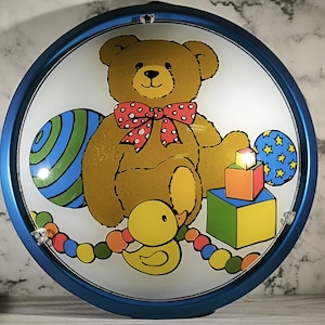 May include: A round, blue-rimmed decorative plate featuring a cartoon teddy bear with a red bow tie, surrounded by toys. Includes a duck, balls, and colorful blocks. The plate is designed with a light background.