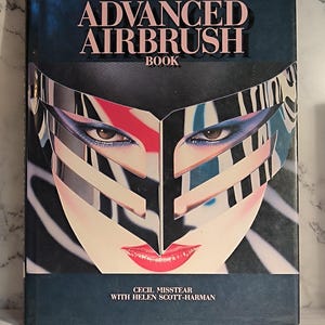以下が含まれることがあります： 「The Advanced Airbrush Book」というタイトルのヴィンテージ本。印象的な表紙のデザインで、銀色のマスクと大胆なメイクアップを施した様式化された顔が、濃い青色の背景に描かれています。著者の名前も表示されています。