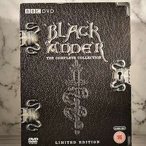 Puede incluir: Un estuche de DVD negro para la serie "Black Adder" con un diseño de serpiente y espada plateadas. El estuche es una edición limitada e incluye 6 discos. El texto "BBC DVD" está en la esquina superior izquierda. El texto "Black Adder" está en una fuente grande y estilizada. El texto "The Complete Collection" está debajo del título. El texto "Limited Edition" está en la parte inferior central de la caja. El texto "6 Disc Set" está en la esquina inferior derecha. El texto "16" está en un círculo rojo en la esquina inferior derecha.
