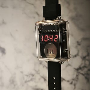 K&ouml;nnte beinhalten: Eine digitale Armbanduhr mit einem transparenten, rechteckigen Geh&auml;use, das die Uhrzeit 10:42 in roten Ziffern anzeigt. Die Uhr hat ein schwarzes Armband mit L&ouml;chern zur Gr&ouml;&szlig;enanpassung. Das Zifferblatt zeigt interne Schaltkreise und einen silbernen Knopf.