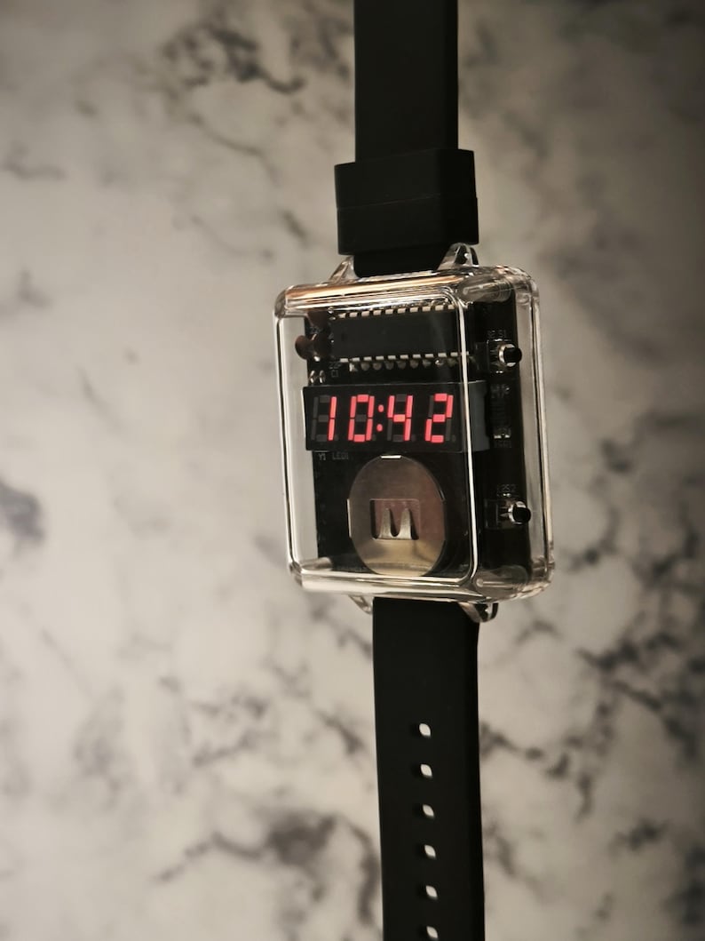 K&ouml;nnte beinhalten: Eine digitale Armbanduhr mit einem transparenten, rechteckigen Geh&auml;use, das die Uhrzeit 10:42 in roten Ziffern anzeigt. Die Uhr hat ein schwarzes Armband mit L&ouml;chern zur Gr&ouml;&szlig;enanpassung. Das Zifferblatt zeigt interne Schaltkreise und einen silbernen Knopf.