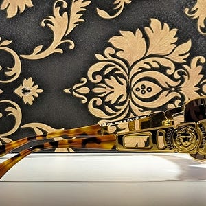 May include: Gold-framed sunglasses with a lion emblem and brown lenses. The temples have a tortoise shell pattern. The sunglasses are set against a black and gold patterned background.