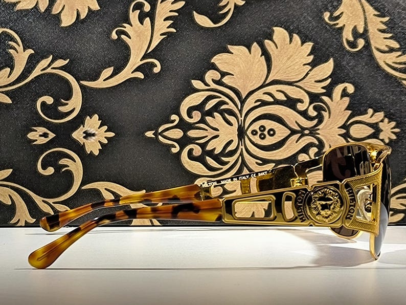 May include: Gold-framed sunglasses with a lion emblem and brown lenses. The temples have a tortoise shell pattern. The sunglasses are set against a black and gold patterned background.
