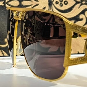 May include: Gold-framed sunglasses with dark lenses. The sunglasses have a decorative design on the frame and the text "HILTON" on the side. The background features a black and gold patterned design.