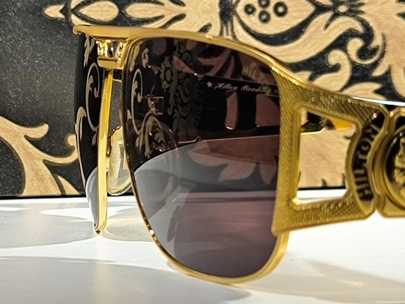 May include: Gold-framed sunglasses with dark lenses. The sunglasses have a decorative design on the frame and the text "HILTON" on the side. The background features a black and gold patterned design.