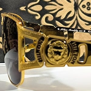 May include: Gold-toned sunglasses with a rectangular frame. The sunglasses feature the word "HILTON" on the side and a lion's head emblem. The lenses are a dark gray color. The text "Killer Piccadilly USA" is also visible.