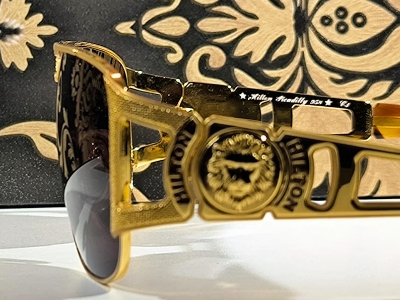 May include: Gold-toned sunglasses with a rectangular frame. The sunglasses feature the word "HILTON" on the side and a lion's head emblem. The lenses are a dark gray color. The text "Killer Piccadilly USA" is also visible.