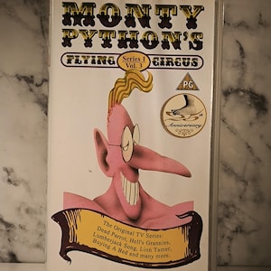 Peut inclure: Une cassette VHS de Monty Python's Flying Circus. La couverture présente un personnage de dessin animé rose avec des lunettes et un long nez. Le texte comprend "Série 1 Vol. 3" et des titres d'épisodes.
