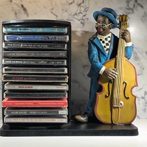 Op de afbeelding: Een zwarte cd-standaard met verschillende muziekalbums, naast een beeldje van een muzikant in een blauw pak die een gele contrabas bespeelt. Het beeldje draagt een blauwe hoed en een bril. De cd-standaard is gevuld met cd's, waaronder "Dark Side of the Moon" van Pink Floyd.