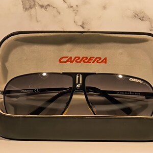 May include: Black Carrera sunglasses with a dark grey gradient lens, resting in an open black case. The case has a light grey interior and the brand name "CARRERA" in red. The sunglasses have a black frame and a silver bridge.