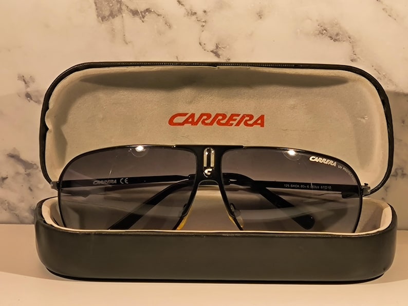 May include: Black Carrera sunglasses with a dark grey gradient lens, resting in an open black case. The case has a light grey interior and the brand name "CARRERA" in red. The sunglasses have a black frame and a silver bridge.