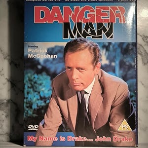 Pode incluir: Conjunto de DVD de "Danger Man: Complete Series One" com Patrick McGoohan. A capa apresenta o título do programa em vermelho e branco sobre um fundo azul e branco. O texto "My Name is Drake... John Drake" também é visível.
