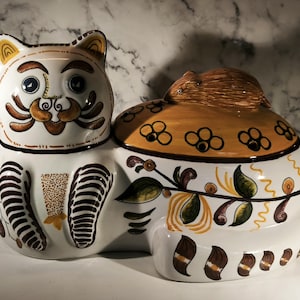 Peut inclure: Un récipient en céramique blanc en forme de chat avec un couvercle, orné de motifs décoratifs bruns et jaunes. Le chat a un visage détaillé avec des yeux bleus et une moustache fantaisiste. Le couvercle est surmonté d'une petite figurine animale brune.