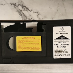 Könnte beinhalten: Schwarzer VHS-C-Kassettenkonverter mit weißem Text und einem gelben Aufkleber mit der Aufschrift "ATTENTION". Der Konverter trägt die Aufschrift "GAWMWAGA VHS-C Cassette Converter" und "VHS-C100 VHS-C Cassette Converter".