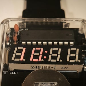 K&ouml;nnte beinhalten: Nahaufnahme eines Digitaldisplays mit rot leuchtenden Zahlen und Doppelpunkten. Das Display befindet sich in einem transparenten Kunststoffgeh&auml;use, das die interne Leiterplatte mit verschiedenen elektronischen Bauteilen freilegt. Der Text "2481BS-1" und "527" ist sichtbar.