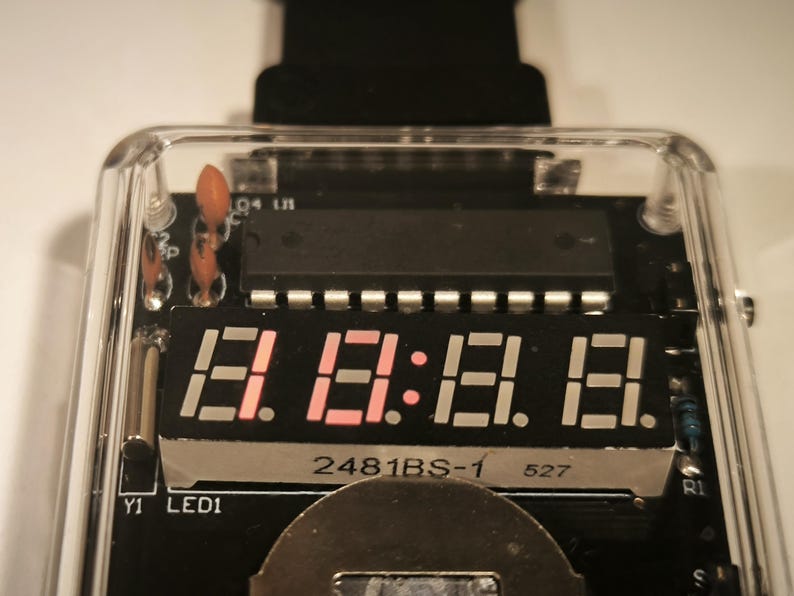 K&ouml;nnte beinhalten: Nahaufnahme eines Digitaldisplays mit rot leuchtenden Zahlen und Doppelpunkten. Das Display befindet sich in einem transparenten Kunststoffgeh&auml;use, das die interne Leiterplatte mit verschiedenen elektronischen Bauteilen freilegt. Der Text "2481BS-1" und "527" ist sichtbar.
