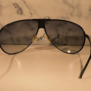 May include: Black aviator sunglasses with dark lenses. The sunglasses have a black frame and temples, with a bridge connecting the lenses. The lenses are a dark gray color. The sunglasses are resting on a white surface.