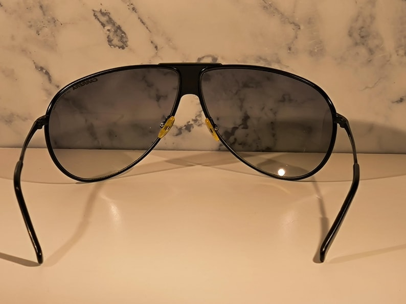 May include: Black aviator sunglasses with dark lenses. The sunglasses have a black frame and temples, with a bridge connecting the lenses. The lenses are a dark gray color. The sunglasses are resting on a white surface.