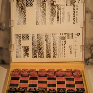 May include: A vintage travel game set with a yellow plastic case. The set includes a checkerboard with red and black squares, red and black game pieces, and instructions. The game pieces are arranged on the board, ready for play.