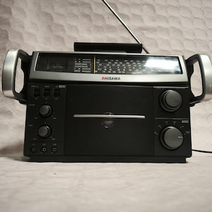 Può includere: Una radio HISWA vintage nera e argento con antenna telescopica e due maniglie argentate. La radio presenta un quadrante di sintonizzazione, manopole di controllo e una griglia per altoparlanti. È un dispositivo elettronico in stile retrò.