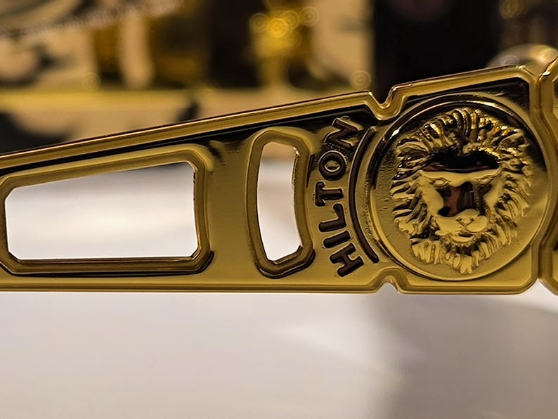 May include: Close-up of a gold-colored sunglass frame. The frame features a rectangular cutout and the word "HILTON" in black lettering. A lion's head emblem is on the side of the frame.