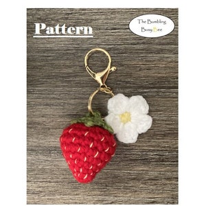 以下が含まれることがあります： 白の編み花の付いた、編み物のイチゴのキーホルダー。キーホルダーは金色の金属製のキーリングに取り付けられています。