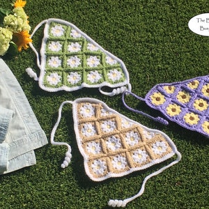 May include: Three crocheted triangle shaped tops with daisy and sunflower designs. The tops are in green, beige, and purple with white accents. The tops are laid out on a green grassy surface.