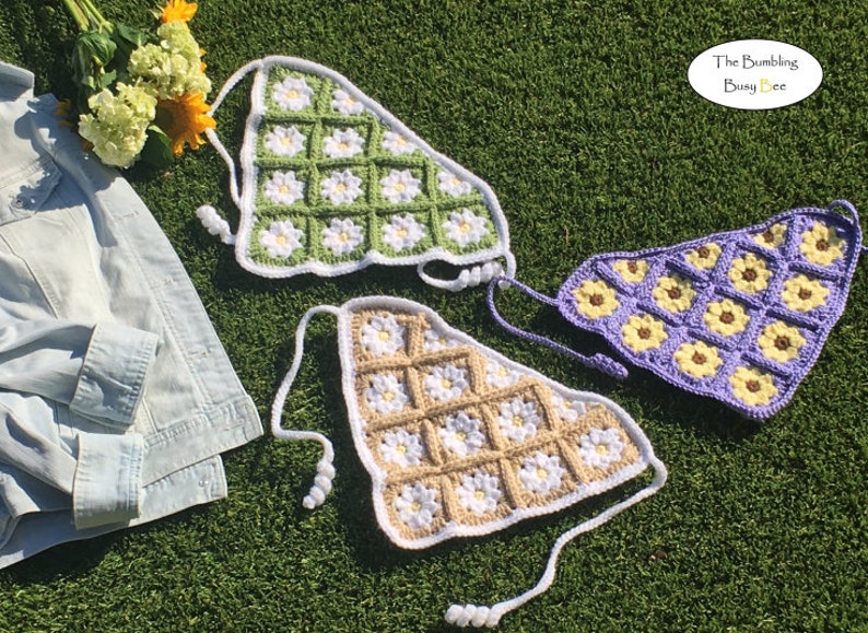 May include: Three crocheted triangle shaped tops with daisy and sunflower designs. The tops are in green, beige, and purple with white accents. The tops are laid out on a green grassy surface.
