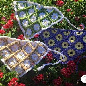May include: Three crocheted triangle tops with daisy flower designs. The tops are in green, purple, and beige with white accents. The tops are laid out on a bed of green leaves and red flowers.