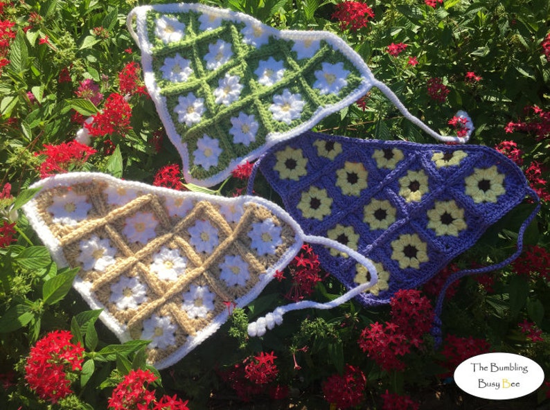 May include: Three crocheted triangle tops with daisy flower designs. The tops are in green, purple, and beige with white accents. The tops are laid out on a bed of green leaves and red flowers.