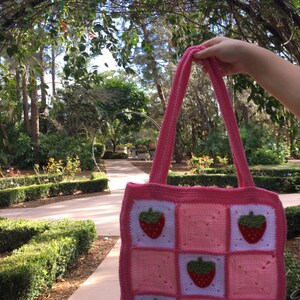 May include: A pink crocheted tote bag with a strawberry design. The bag has a white background with pink squares and strawberries in each square.