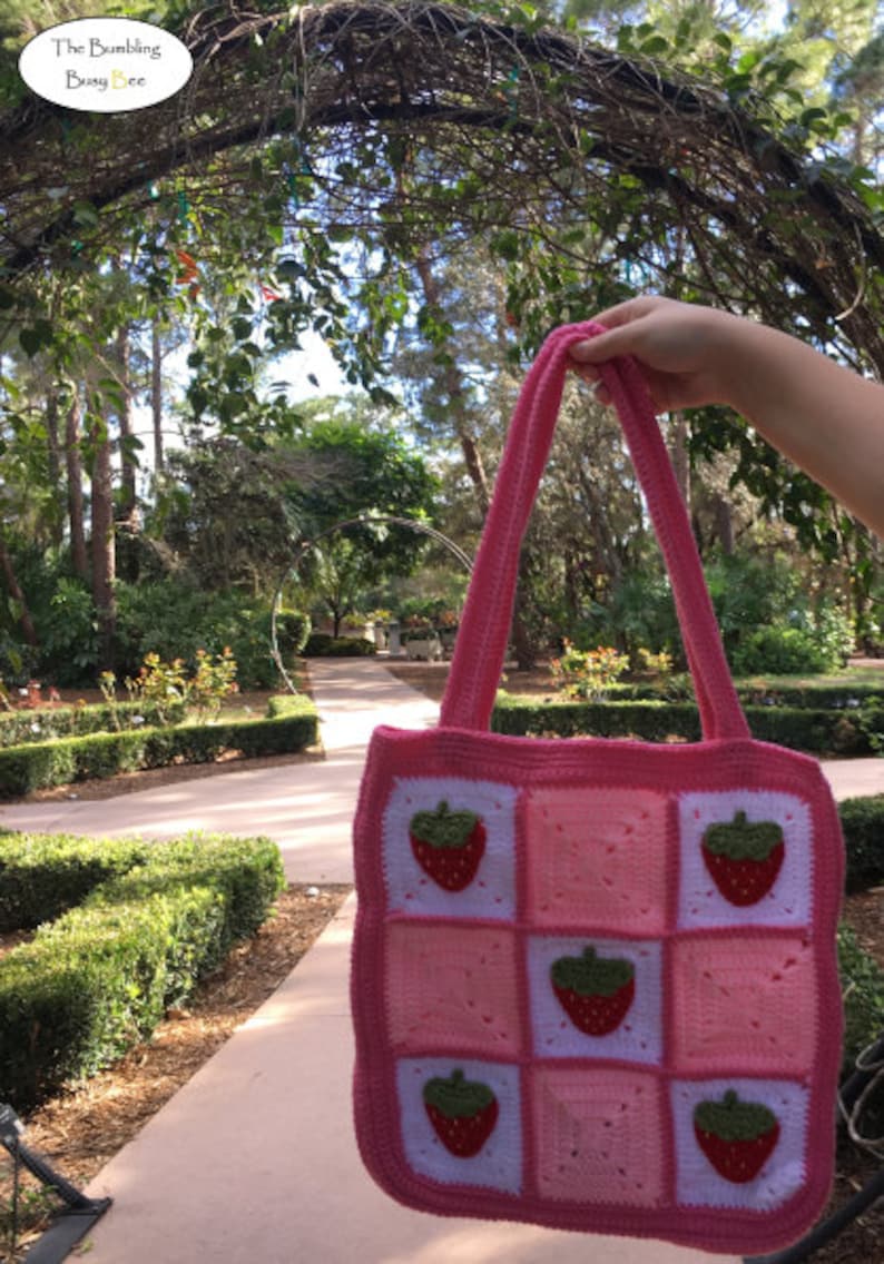 May include: A pink crocheted tote bag with a strawberry design. The bag has a white background with pink squares and strawberries in each square.