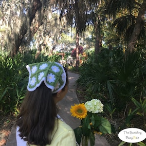 May include: A woman with long brown hair is wearing a green and white crocheted headband with a daisy pattern. She is holding a bouquet of white hydrangeas and a yellow sunflower. The background is a lush green garden with a path.