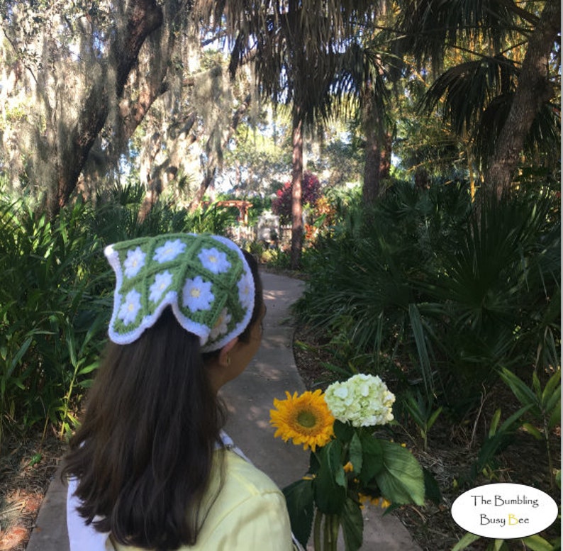 May include: A woman with long brown hair is wearing a green and white crocheted headband with a daisy pattern. She is holding a bouquet of white hydrangeas and a yellow sunflower. The background is a lush green garden with a path.