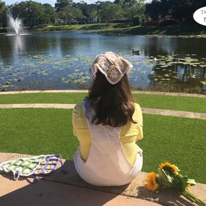 May include: A woman wearing a white crocheted hat with a yellow flower design sits on a stone ledge overlooking a pond with lily pads. She is wearing a yellow shirt and white overalls. There are sunflowers on the ground in front of her.