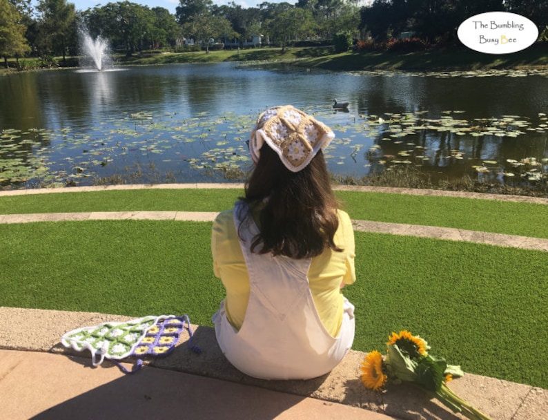 May include: A woman wearing a white crocheted hat with a yellow flower design sits on a stone ledge overlooking a pond with lily pads. She is wearing a yellow shirt and white overalls. There are sunflowers on the ground in front of her.