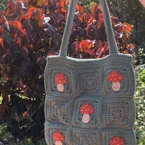 May include: Handmade crochet tote bag in a sage green color, featuring nine square panels. Each panel is adorned with a mushroom applique in shades of red, white, and peach. The bag has long handles and the text "The Bumbling Busy Bee" in a white oval.