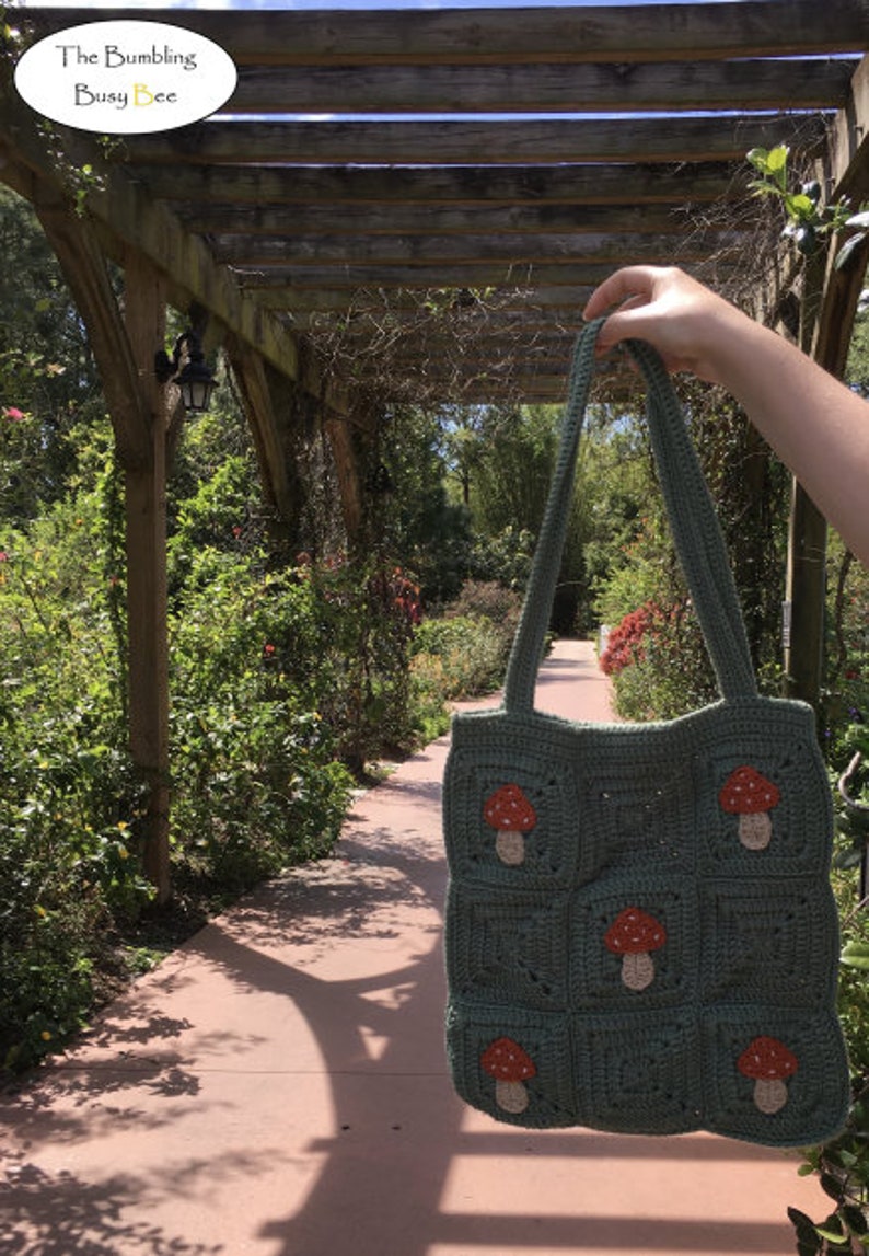 May include: A handmade green crochet tote bag with a mushroom design. The bag features multiple squares, each adorned with an embroidered mushroom. The bag is held up in front of a garden path. The text "The Bumbling Busy Bee" is in the top left corner.
