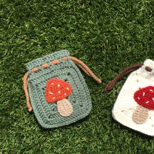 May include: Two crocheted drawstring bags with mushroom appliqu&eacute;s. One bag is sage green with an orange and beige mushroom, the other is cream with a red and beige mushroom. The bags are on a green grassy surface.