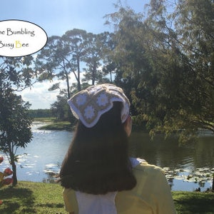 May include: A white and beige crocheted headband with a daisy flower design. The headband is worn by a person with long brown hair, standing in front of a lake and trees.