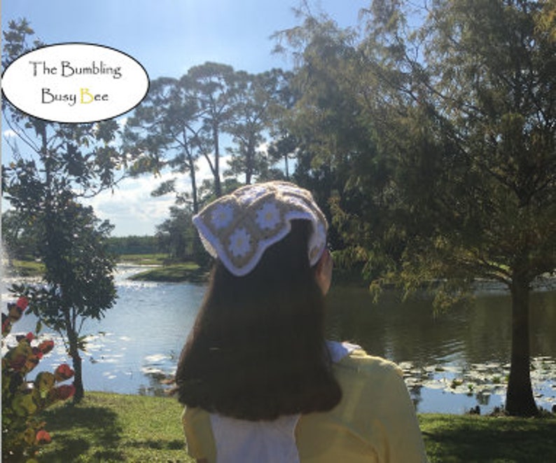 May include: A white and beige crocheted headband with a daisy flower design. The headband is worn by a person with long brown hair, standing in front of a lake and trees.