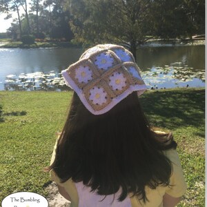 May include: A white and beige crochet hat with a daisy pattern. The hat is worn by a person with long brown hair, standing in front of a pond.