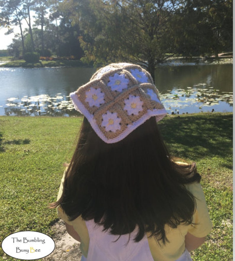 May include: A white and beige crochet hat with a daisy pattern. The hat is worn by a person with long brown hair, standing in front of a pond.