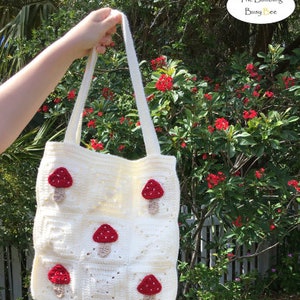May include: Cream-colored crochet tote bag with red mushroom appliqu&eacute;s. The bag features a grid pattern and long straps. The text "The Bumbling Busy Bee" is in the upper right corner. The bag is held up against a background of green foliage and red flowers.