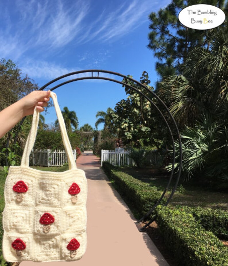 May include: Cream-colored crochet tote bag with red mushroom appliqu&eacute;s. The bag features a square pattern with nine mushroom designs. The bag is held up against a background of a sunny day with a blue sky and a garden path.
