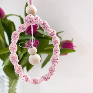 Peut inclure: Ornement en macramé rose avec un design circulaire, comprenant une perle en bois en haut et deux perles en bois suspendues au centre. L'ornement est suspendu devant un arrière-plan flou de tulipes roses et de feuilles vertes.