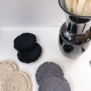 Könnte beinhalten: Eine Auswahl an Makramee-Untersetzern in Schwarz, Beige und Grau. Jeder Untersetzer hat ein Spiraldesign mit einem Fransenrand. Die Untersetzer sind auf einer weißen Oberfläche angeordnet, mit einer schwarzen Vase mit getrockneten Blumen im Hintergrund. Durchmesser ca. 10 cm.