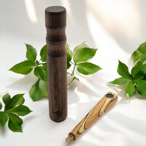May include: A wooden smoking pipe with a brown and black wood grain pattern. The pipe is engraved with the words "Calumet Spiritpipes". The pipe is sitting on a white surface with green leaves in the background.
